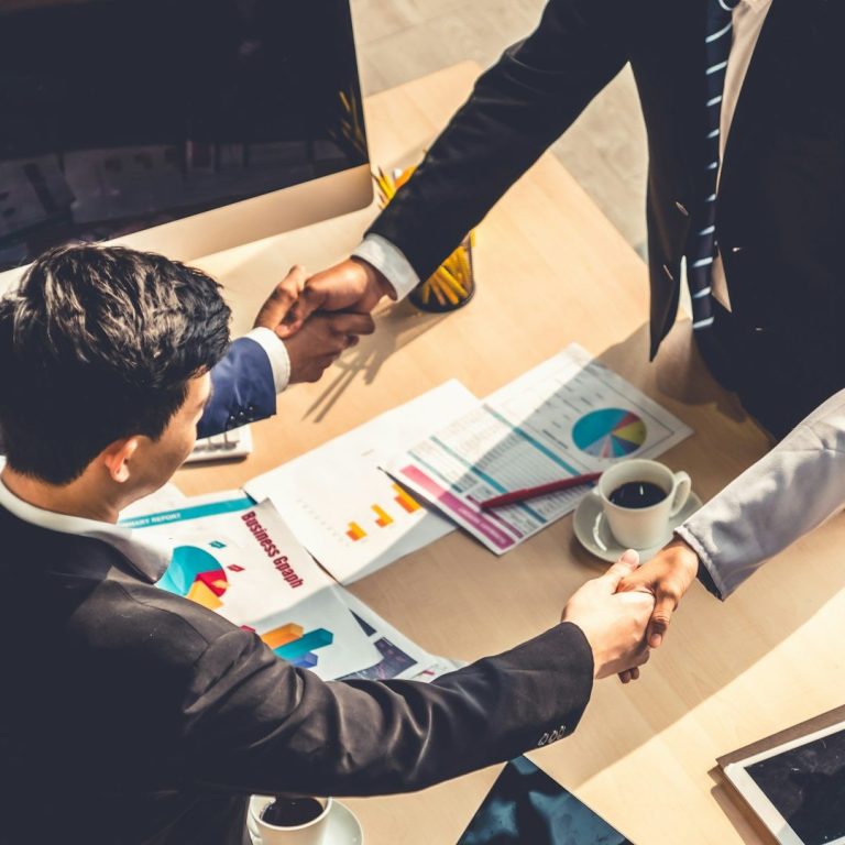 Geschäftsleute schütteln sich die Hände über einem Tisch mit Diagrammen und einer Tasse Kaffee.